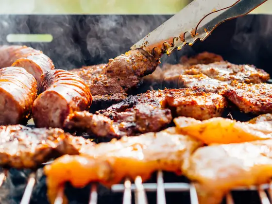 Una pinza gira la carne sulla griglia fumante, accanto a salsicce e petti di pollo