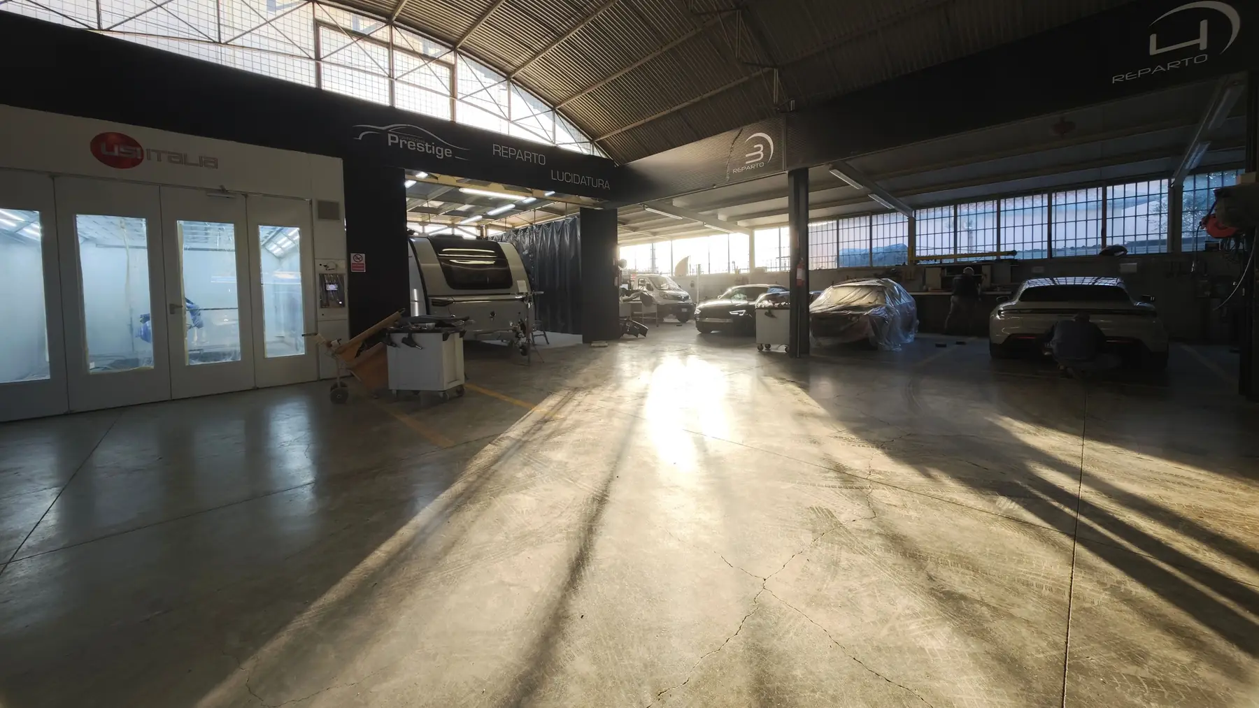 L'officina vista dal fondo: i forni a sinistra, le auto in lavorazione e la luce del mattino sul pavimento