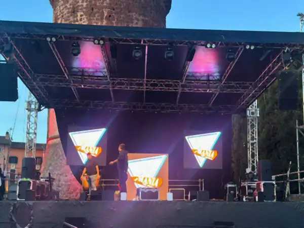 Palco con il tetto a traliccio e tre schermi LED a triangolo, davanti a una torre antica