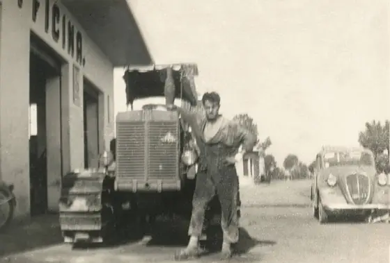 Foto d'epoca: un uomo in tuta accanto a un trattore cingolato, davanti all'officina