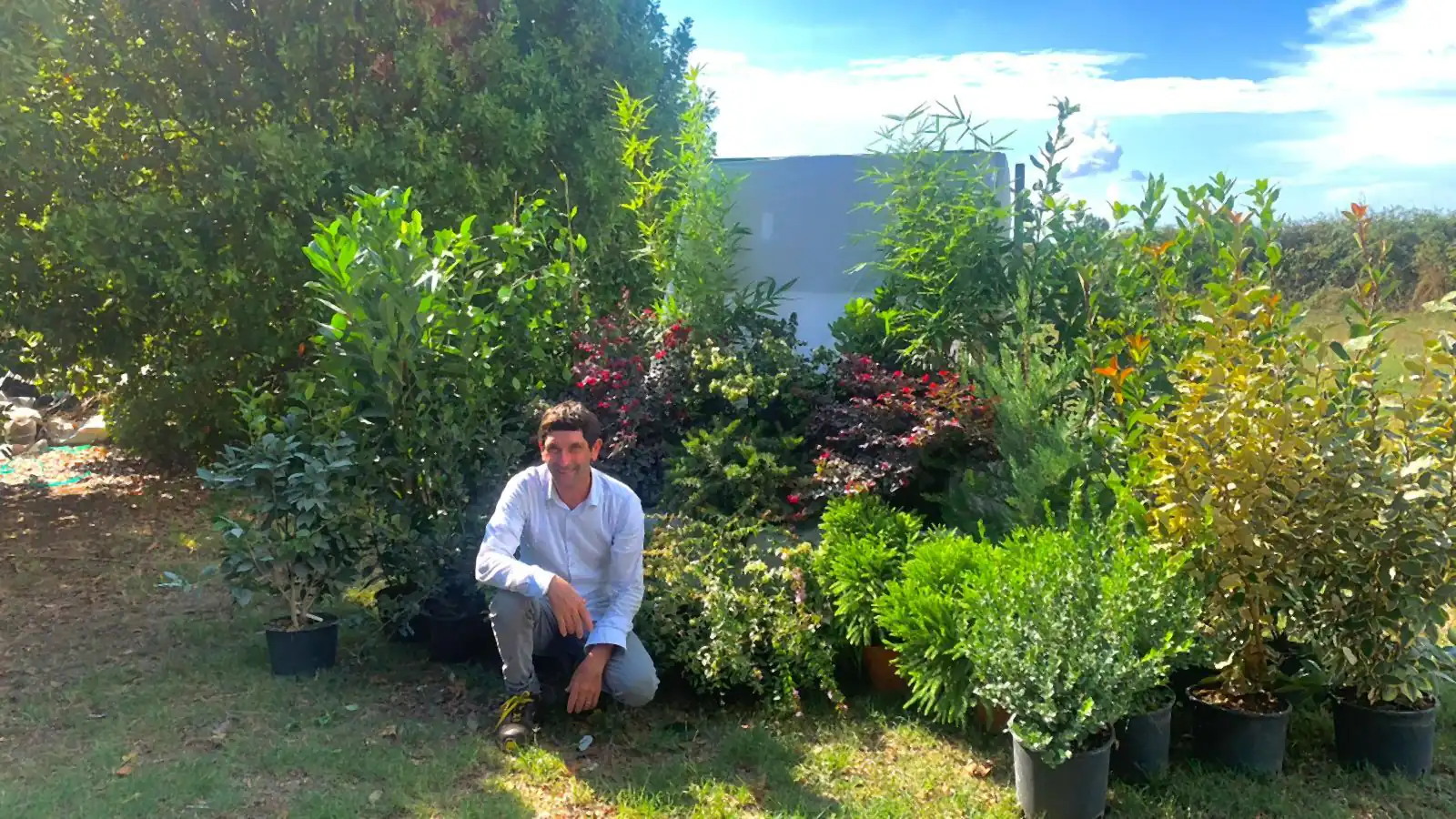 Un uomo accovacciato sul prato fra arbusti e piante fiorite in vaso