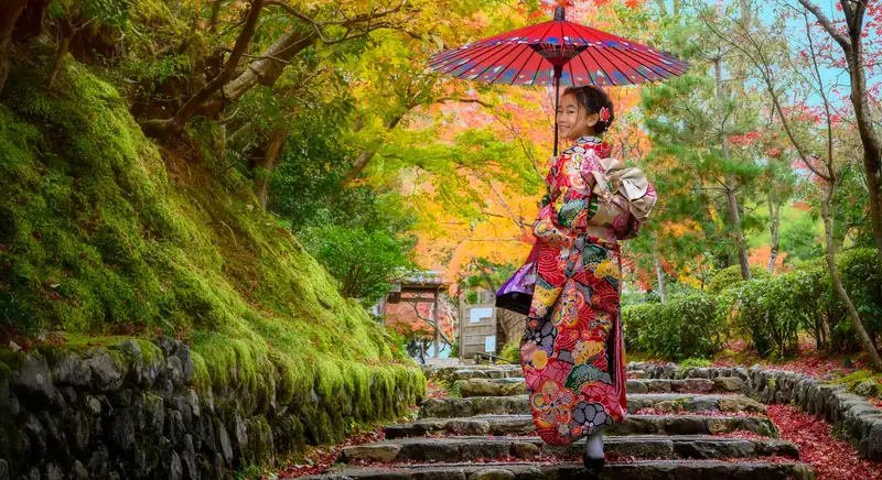 Una bambina in kimono con l’ombrello rosso sale una scalinata di pietra fra gli aceri d’autunno, voltandosi a sorridere
