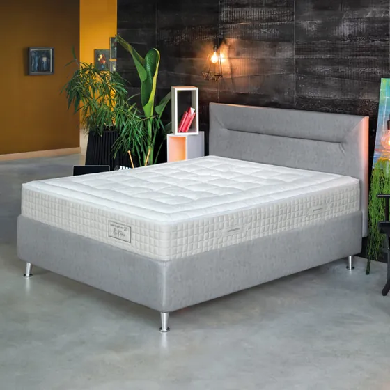 Letto con giroletto grigio e materasso Permaflex trapuntato, in una stanza con piante e una parete scura