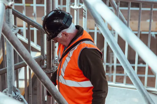 In cantiere, un uomo col casco e il giubbotto ad alta visibilità passa fra i tubi del ponteggio